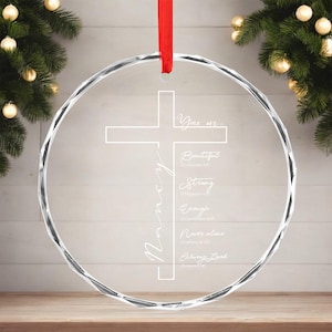 May include: A clear, circular Christmas ornament featuring a cross design and the name "Nancy" in script. The ornament has the words "You are... Beautiful, Strong, Enough, Never alone, Always Loved." A red ribbon is attached for hanging.
