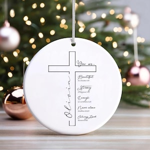 May include: A white ceramic Christmas ornament featuring a cross design and the name "Olivia" in cursive. The ornament includes the phrases "You are... Beautiful, Strong, Enough, Never alone, Always Loved."