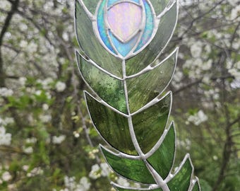 May include: A stained glass peacock feather with a green and blue iridescent design. The feather is hanging from a white ribbon.