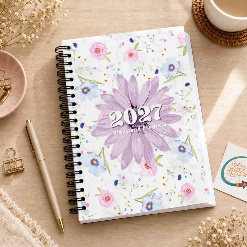 May include: A spiral-bound planner with a white cover featuring a pattern of small pink, blue, and yellow flowers. A large, central purple flower graphic displays the text 2027 Plans and Progress in white lettering. The planner has a black wire coil.