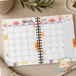 May include: A spiral-bound 2027 calendar open to the month of January. The pages feature a white grid layout with black text for days of the week and dates, decorated with watercolour-style orange, yellow, and pink floral illustrations.