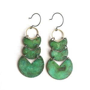 May include: A pair of green and gold earrings with three tiers of crescent-shaped metal pieces. The earrings have a simple, modern design.
