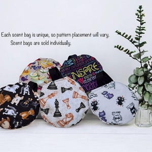 May include: A collection of round scent bags with various patterns, including dogs, chess pieces, and abstract designs. One bag features the word "INSPIRE". The bags have black handles and are displayed on a white surface. The text says: "Each scent bag is unique, so pattern placement will vary."