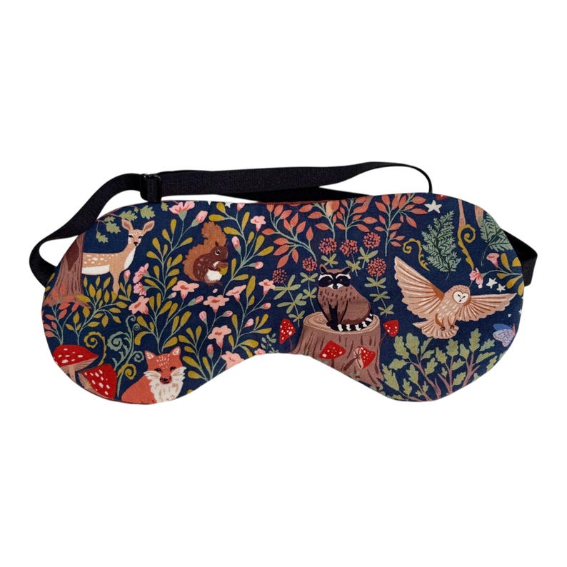 K&ouml;nnte beinhalten: Eine marineblaue Schlafmaske mit einem Waldmotiv, das ein Reh, ein Eichh&ouml;rnchen, einen Fuchs, eine Eule und einen Waschb&auml;ren zeigt. Die Maske hat ein schwarzes Gummiband und ist mit Blumen, Pilzen und Bl&auml;ttern verziert.