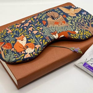 K&ouml;nnte beinhalten: Eine marineblaue Schlafmaske mit einem Waldmuster aus Fuchs und Eule, die auf einem braunen Notizbuch liegt. Die Maske zeigt orange F&uuml;chse, rote Pilze und einen Baumstumpf. Ein kleiner Lavendelbeutel ist ebenfalls vorhanden.