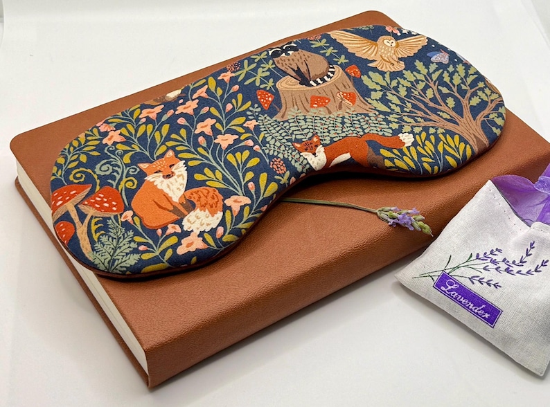 K&ouml;nnte beinhalten: Eine marineblaue Schlafmaske mit einem Waldmuster aus Fuchs und Eule, die auf einem braunen Notizbuch liegt. Die Maske zeigt orange F&uuml;chse, rote Pilze und einen Baumstumpf. Ein kleiner Lavendelbeutel ist ebenfalls vorhanden.