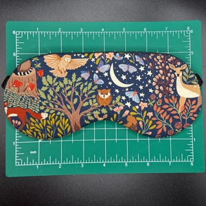 K&ouml;nnte beinhalten: Eine marineblaue Schlafmaske mit einer skurrilen Waldszene mit einer Eule, einem Hirsch, Schmetterlingen und einem Halbmond. Die Maske ist etwa 20 cm lang und 7,6 cm hoch, mit einem schwarzen Gummiband.