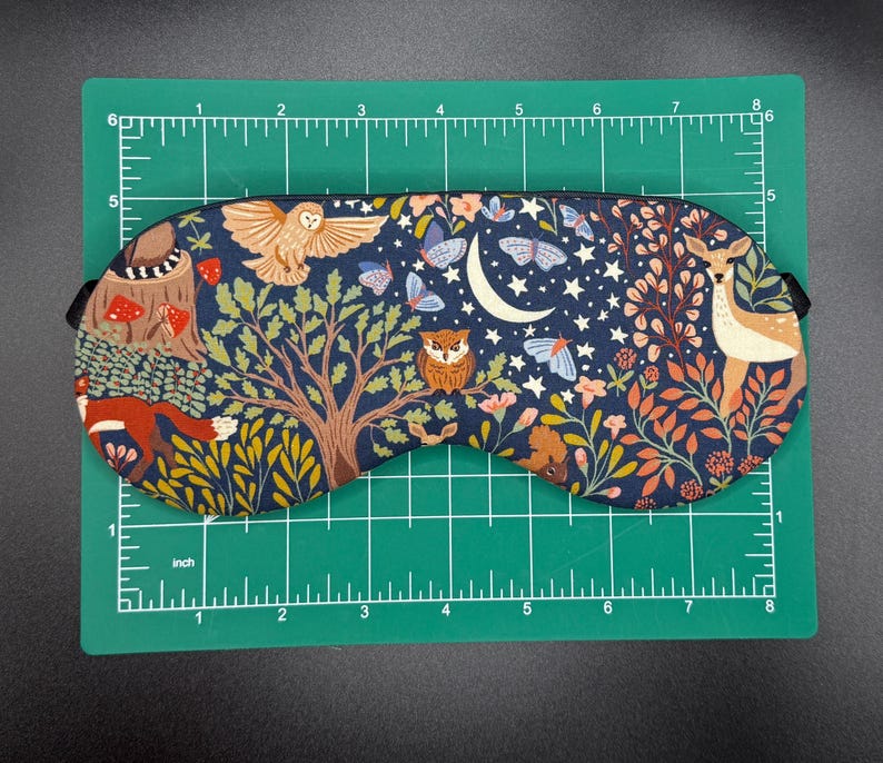 K&ouml;nnte beinhalten: Eine marineblaue Schlafmaske mit einer skurrilen Waldszene mit einer Eule, einem Hirsch, Schmetterlingen und einem Halbmond. Die Maske ist etwa 20 cm lang und 7,6 cm hoch, mit einem schwarzen Gummiband.