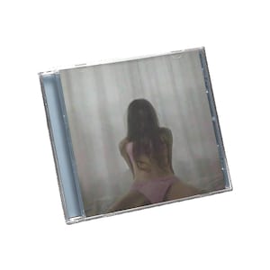 Puede incluir: Una caja de CD transparente con una fotografía en su interior. La fotografía muestra a una persona con un traje de baño rosa. La persona está sentada en una cama de espaldas al espectador. El fondo es una cortina borrosa de color claro.