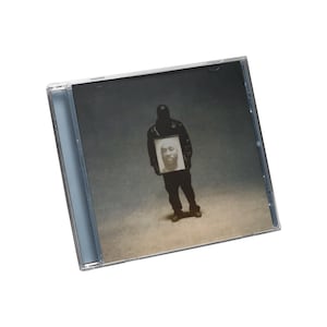 May include: A CD case featuring a person in a black outfit holding a framed portrait. The background is a muted, sepia-toned scene. The CD case is clear with a gray-toned cover.