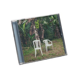 Puede incluir: Una caja de CD con una foto de dos sillas de plástico blancas sobre hierba. El fondo muestra follaje verde, plataneros y un racimo de plátanos. La escena evoca un entorno tropical o al aire libre.