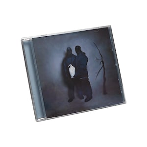 May include: A CD case featuring a dark, moody album cover. The artwork depicts two figures in dark clothing, one holding a white bird. A bare tree stands in the background, all set against a gray backdrop.