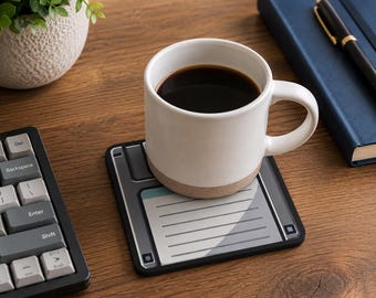 May include: A square coaster designed to look like a vintage floppy disk with a grey and black color scheme, featuring a central label area with horizontal lines. A white ceramic mug filled with black coffee sits on top of the coaster.