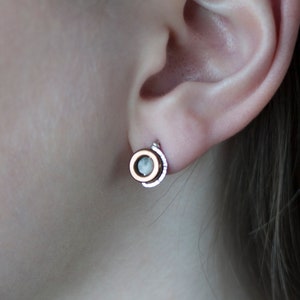 Könnte beinhalten: Ein Paar kleine, runde Ohrringe mit einem silbernen und goldenen Design. Die Ohrringe haben einen kleinen, klaren Stein in der Mitte.