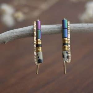 Pode incluir: Um par de brincos de ouro com um design geométrico. Os brincos apresentam uma mistura de contas pretas, brancas e iridescentes de roxo e azul.