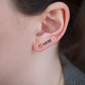 Può includere: Primo piano di un orecchino oro e nero. L'orecchino ha un piccolo cerchio d'oro in basso, una barra nera e oro al centro e un cerchio d'oro in alto. L'orecchino è nell'orecchio di una persona.