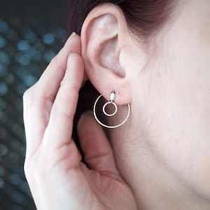 May include: A pair of silver hoop earrings with a smaller circle inside the larger circle. The earrings are worn in a person's ear.