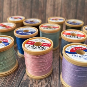 May include: A selection of colourful cotton thread spools, including shades of green, blue, pink, and lilac. Each spool, branded "Tootal", is labelled with the thread type and length of 100 m. The spools are arranged on a wooden surface.