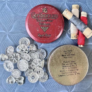 May include: A collection of vintage sewing supplies, including clear glass buttons, spools of thread, and two metal tins. One tin is red with "Caribonum Typewriter Ribbon" text. The other tin is gold with text. The background is a blue floral pattern.