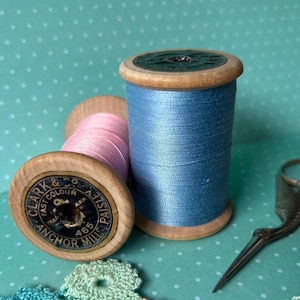 Könnte beinhalten: Zwei Holzspulen mit Garn, eine blaue und eine rosa, mit einer silbernen Schere auf einem türkisfarbenen Hintergrund. Die blaue Spule hat den Text "Clark & Co. Fast Colour Anchor Mills 465 Paisley" auf der Seite aufgedruckt.