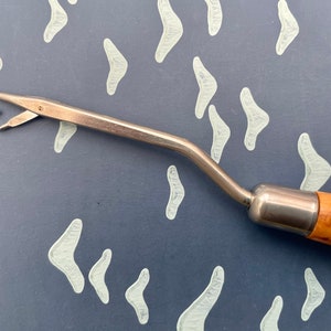 May include: A vintage rug hooking tool with a wooden handle and a metal hook. The tool has a curved design and a small blade near the hook. The handle is made of light-coloured wood.