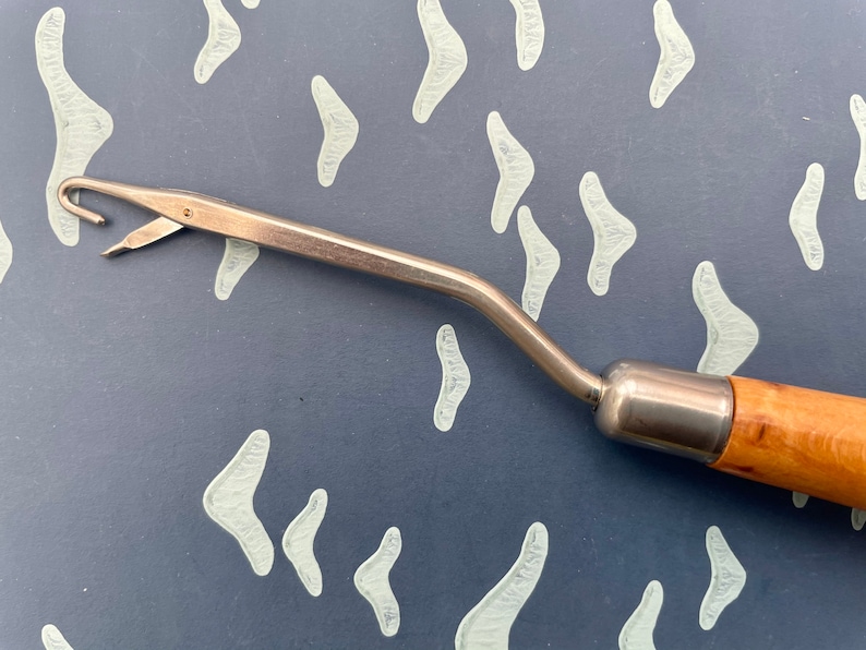 May include: A vintage rug hooking tool with a wooden handle and a metal hook. The tool has a curved design and a small blade near the hook. The handle is made of light-coloured wood.