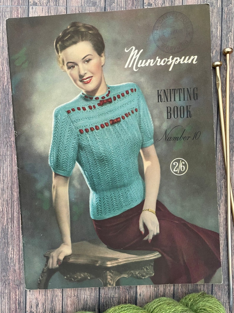 Munrospun knitting book number 10, showing a woman modelling a turquoise handknitted lace jumper with red ribbon threaded through top in two bands. She is perched on a side table. The knitting book is laid flat on table and accessorised with some gold knitting needles.