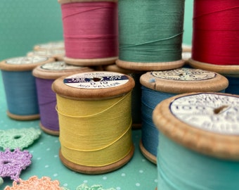 May include: A collection of wooden spools of thread in various colours, including pink, green, blue, yellow, and purple. The spools are stacked on top of each other and some are lying on a green surface. The spools have labels with text that is partially visible.