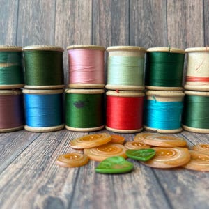 May include: Twelve wooden spools of thread in various colors including teal, dark green, pink, light green, red, blue, and cream, arranged in two rows. Scattered in front are several round, translucent amber buttons and two green leaf-shaped buttons.