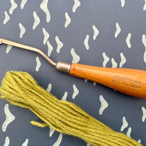 May include: A rug hooking tool with a wooden handle, labelled "neadicut MADE IN ENGLAND", and a bundle of olive green yarn. The tool has a metal hook and a curved design. The yarn is tightly wound.