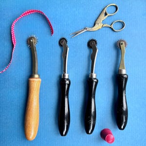 May include: Four leather crafting tools with wooden and black handles. The tools have a wheel on the end of the handle. A pair of gold scissors with a bird design is also in the image.