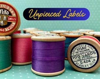 May include: A collection of vintage wooden spools wrapped in colourful mercerised cotton thread. The spools feature circular paper labels with text including J. Dewhurst & Sons, Sylko, 100 Yards, and Scarlet Glow. A blue graphic reads Unpierced Labels.
