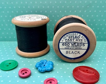 May include: Two wooden spools of black thread labeled "SYLKO FAST DYE" and "650 YARDS" sit next to colorful buttons. The buttons are in shades of green, pink, and blue, with various shapes and designs. The thread is mercerized cotton.