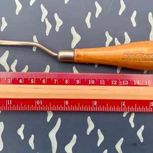 May include: A rug hooking tool with a wooden handle and a metal hook. The handle is light brown with the text "needlecut MADE IN ENGLAND". A ruler is placed below the tool, showing measurements in centimetres.