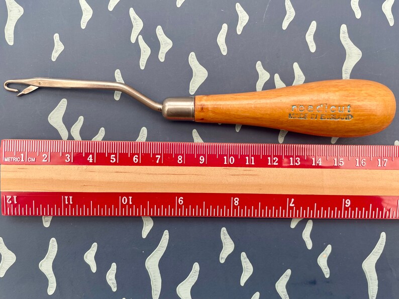 May include: A rug hooking tool with a wooden handle and a metal hook. The handle is light brown with the text "needlecut MADE IN ENGLAND". A ruler is placed below the tool, showing measurements in centimetres.