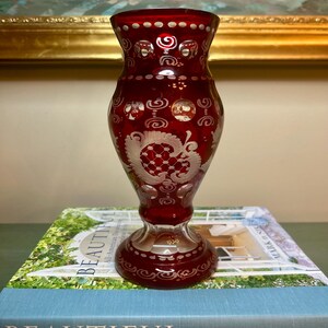 May include: A ruby red glass vase featuring intricate cut-glass patterns, including circular motifs and detailed floral engravings. The vase has a flared rim, a tapered neck, and a wide, circular base, showcasing a clear glass pedestal foot.