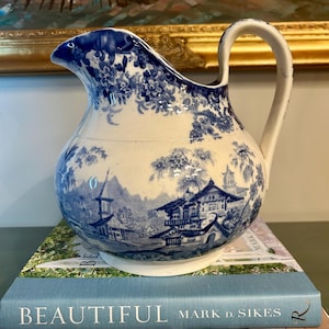 May include: A blue and white ceramic pitcher with a handle. The pitcher features a detailed landscape design and floral patterns. It is sitting on top of a stack of books with the word "BEAUTIFUL" visible.