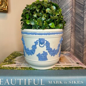 May include: A light blue and white ceramic planter featuring raised neoclassical relief designs of lion heads, floral swags, and figures. The planter contains a dense, rounded artificial green shrub with small, heart-shaped leaves.