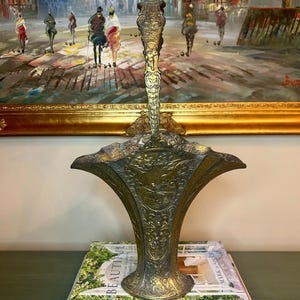 May include: An ornate, antique-style brass basket with a tall, arched handle and a flared, scalloped rim. The surface features intricate embossed floral and scrollwork patterns, with a central medallion depicting a figure in a landscape scene.
