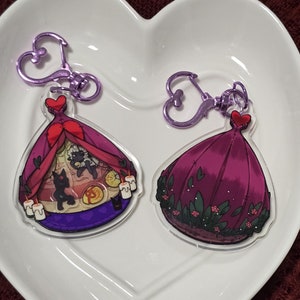 May include: Two purple and pink themed acrylic charms with metal keychains. The charms are shaped like tents with different designs. One charm features a tent with a red roof and a black cat inside. The other charm features a tent with a purple roof and a floral design.