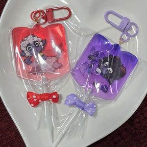 May include: Two colourful lollipop keychains featuring cartoon characters. The red lollipop has a white sheep with horns and a red cape. The purple lollipop has a black sheep with horns and a purple cape. Both lollipops have white sticks and are tied with a bow.