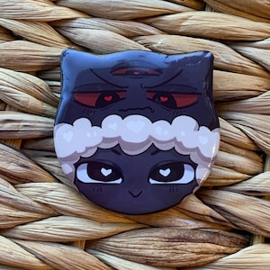May include: A dark blue cat-shaped button with a stylized design. The cat has red eyes and a third eye, with white fluffy accents and heart-shaped pupils. The button is set against a woven background.