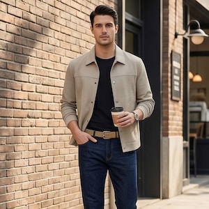 May include: A person wearing a beige jacket over a black t-shirt, dark blue jeans, and white sneakers. The person is holding a coffee cup and wearing a watch. A light brown belt is visible. The background features a brick wall and a storefront.