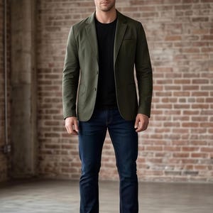 A man wearing a green blazer, black shirt, and dark blue jeans stands in front of a brick wall.