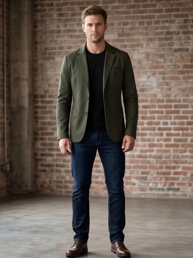 A man wearing a green blazer, black shirt, and dark blue jeans stands in front of a brick wall.