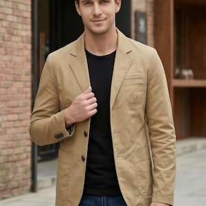 A man wearing a tan blazer jacket, black shirt, and blue jeans stands in front of a brick building.
