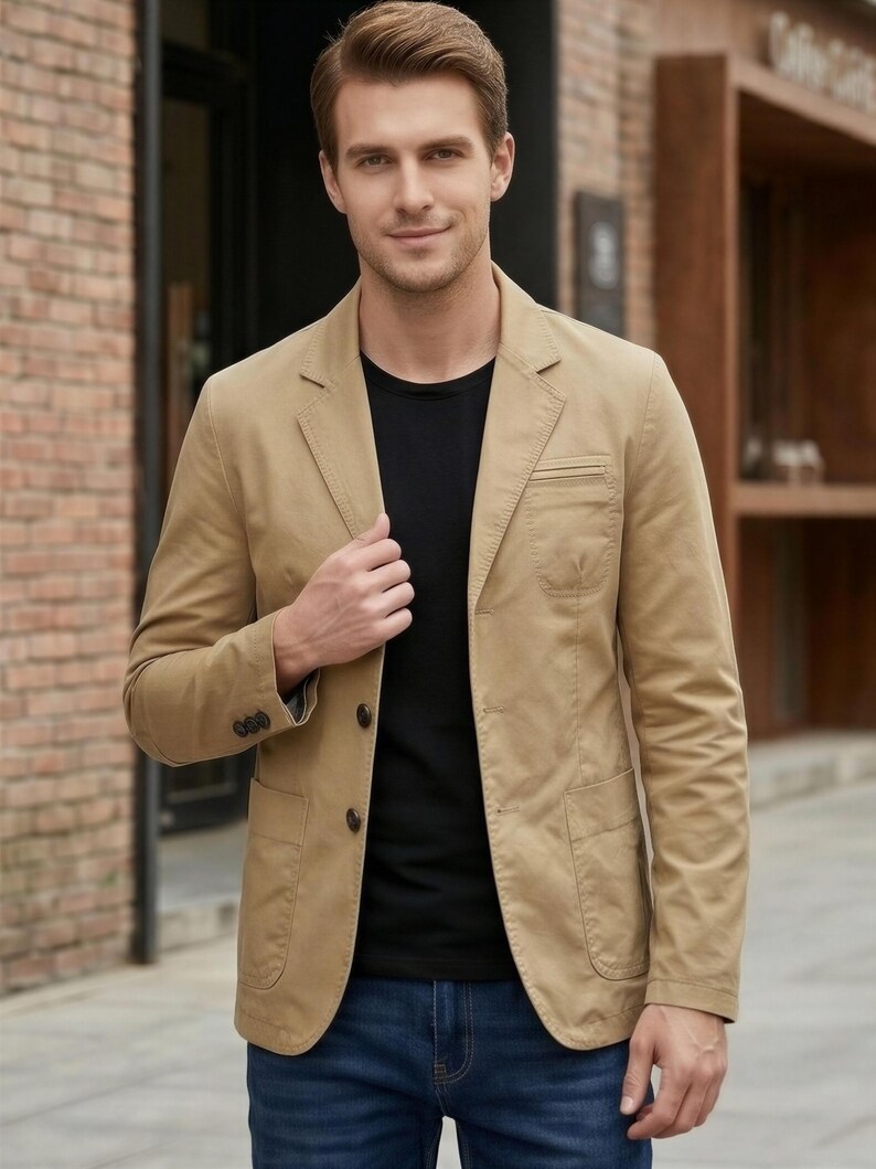 A man wearing a tan blazer jacket, black shirt, and blue jeans stands in front of a brick building.