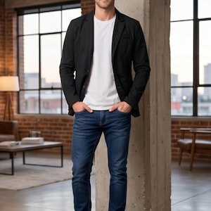 A man wearing a black blazer, white t-shirt, and blue jeans stands in a room with large windows, looking directly at the camera.