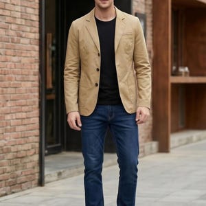 A man wearing a tan blazer, black shirt, and blue jeans stands in front of a brick building.
