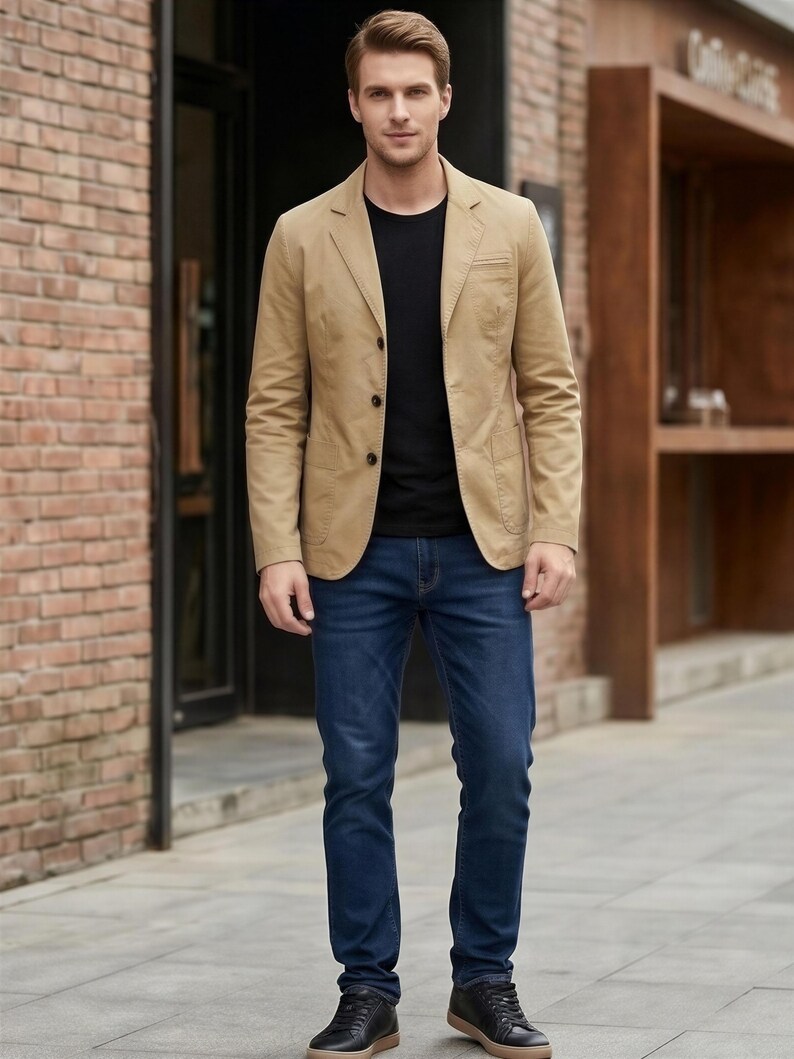 A man wearing a tan blazer, black shirt, and blue jeans stands in front of a brick building.